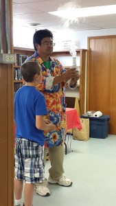 presenter in colorful shirt holds beaker with smoke or vapor coming out of top while child in blue shirt stands nearby